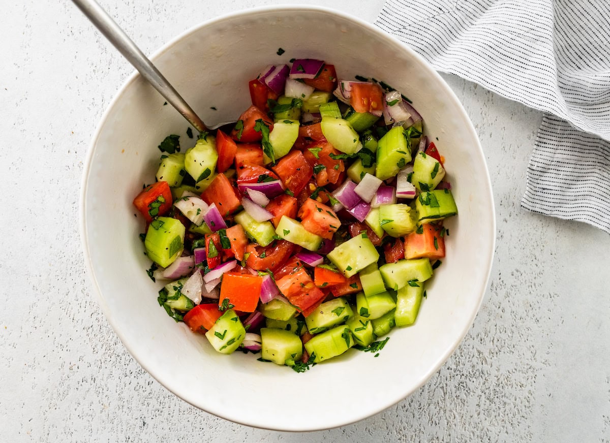 Chopped cucumber, red onion, tomatoes and fresh parsley mixed together in a bowl with lemon juice and olive oil. There is a silver spoon in the bowl.