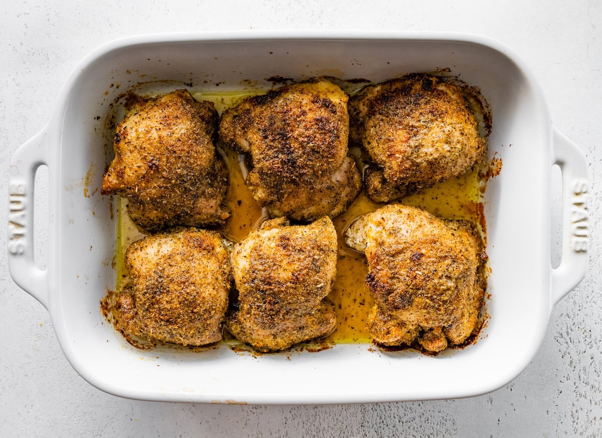 Six baked chicken thighs in a white baking dish coated in the Greek chicken seasoning mixture.