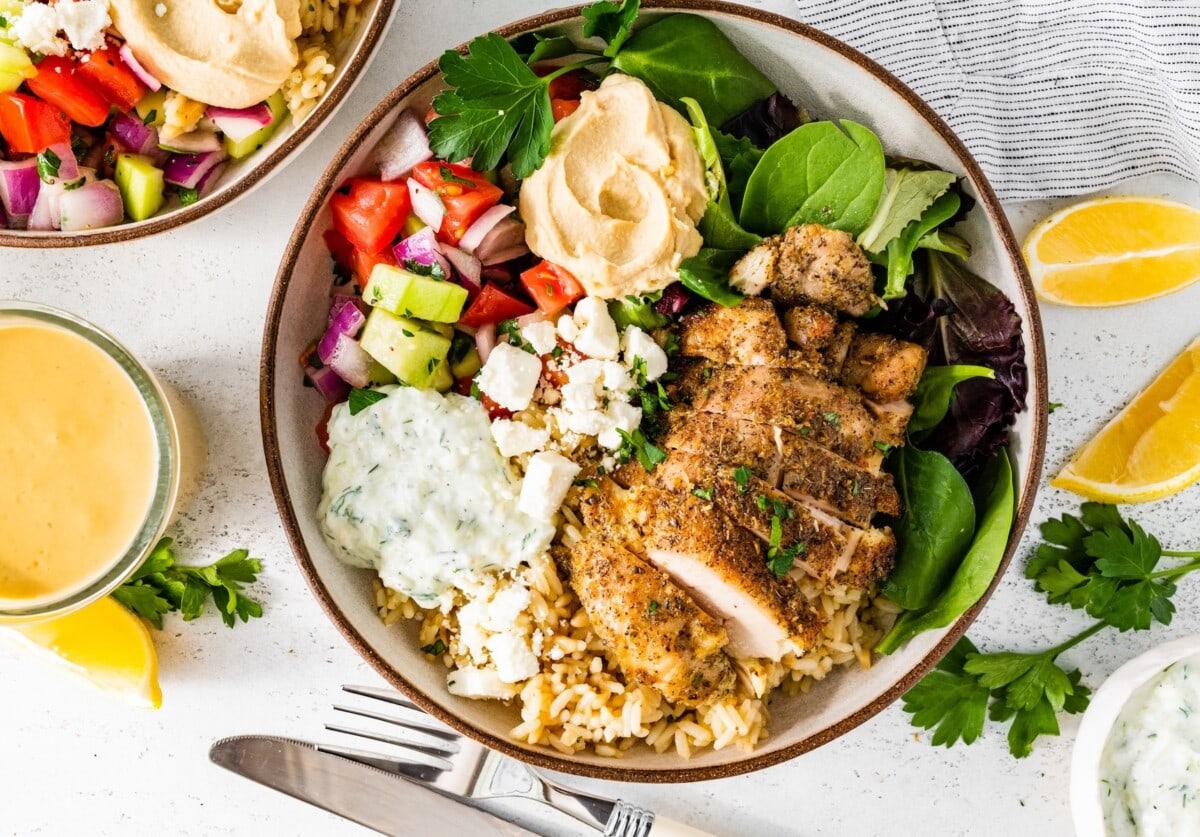 A serving of spiced Greek chicken over a bed of greens, a dollop of hummus, tzatziki, and a cucumber tomato salad.