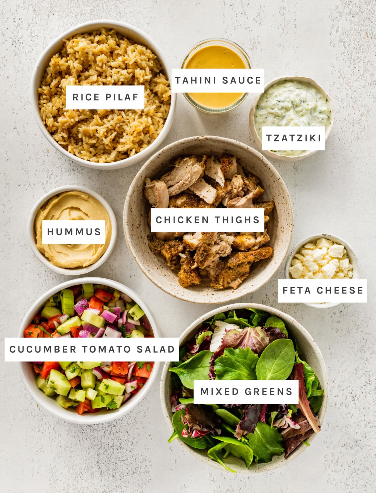 Ingredients measured out to make Greek Chicken Bowls: Rice Pilaf, Parsley, Mixed Greens, Feta Cheese, Tzatziki, Hummus, Chicken Thighs, Tahini Sauce,