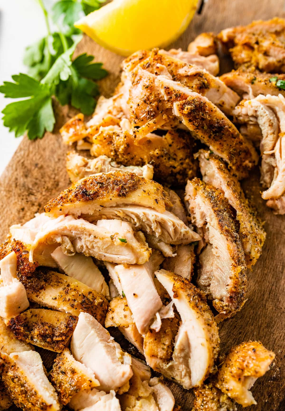 Sliced Greek chicken thighs on a wooden cutting board. There are lemon slices and fresh parsley on the cutting board as well.