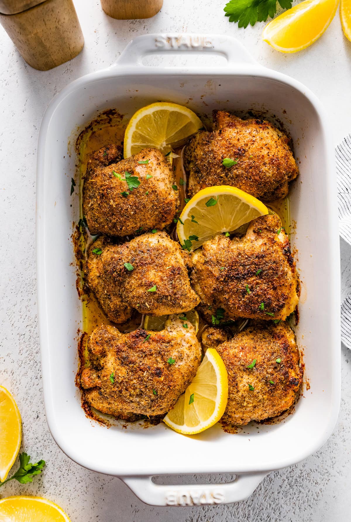 Six baked Greek chicken thighs in a white baking dish. The chicken is topped with fresh parsley and lemon slices.