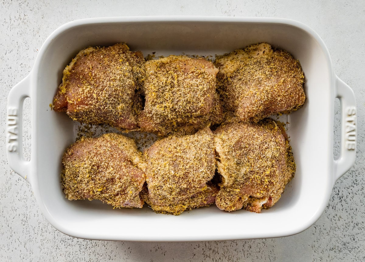 Six raw chicken thighs in a white baking dish. The chicken is coated in a seasoning mixture.
