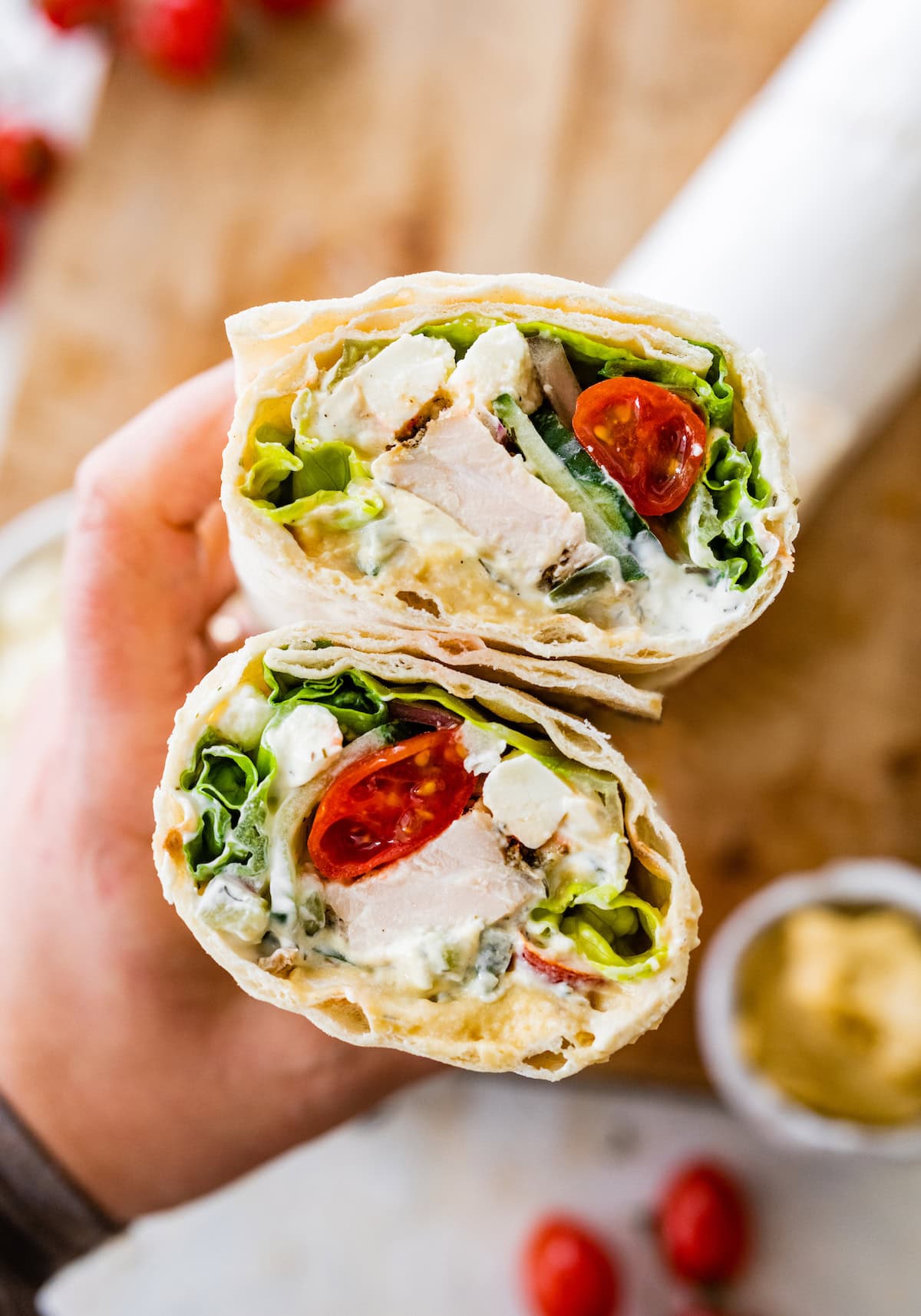 A woman's hand holding a Greek chicken wrap that has been cut in half. You can see the tomatoes, chicken, lettuce and sauce inside the wrap.