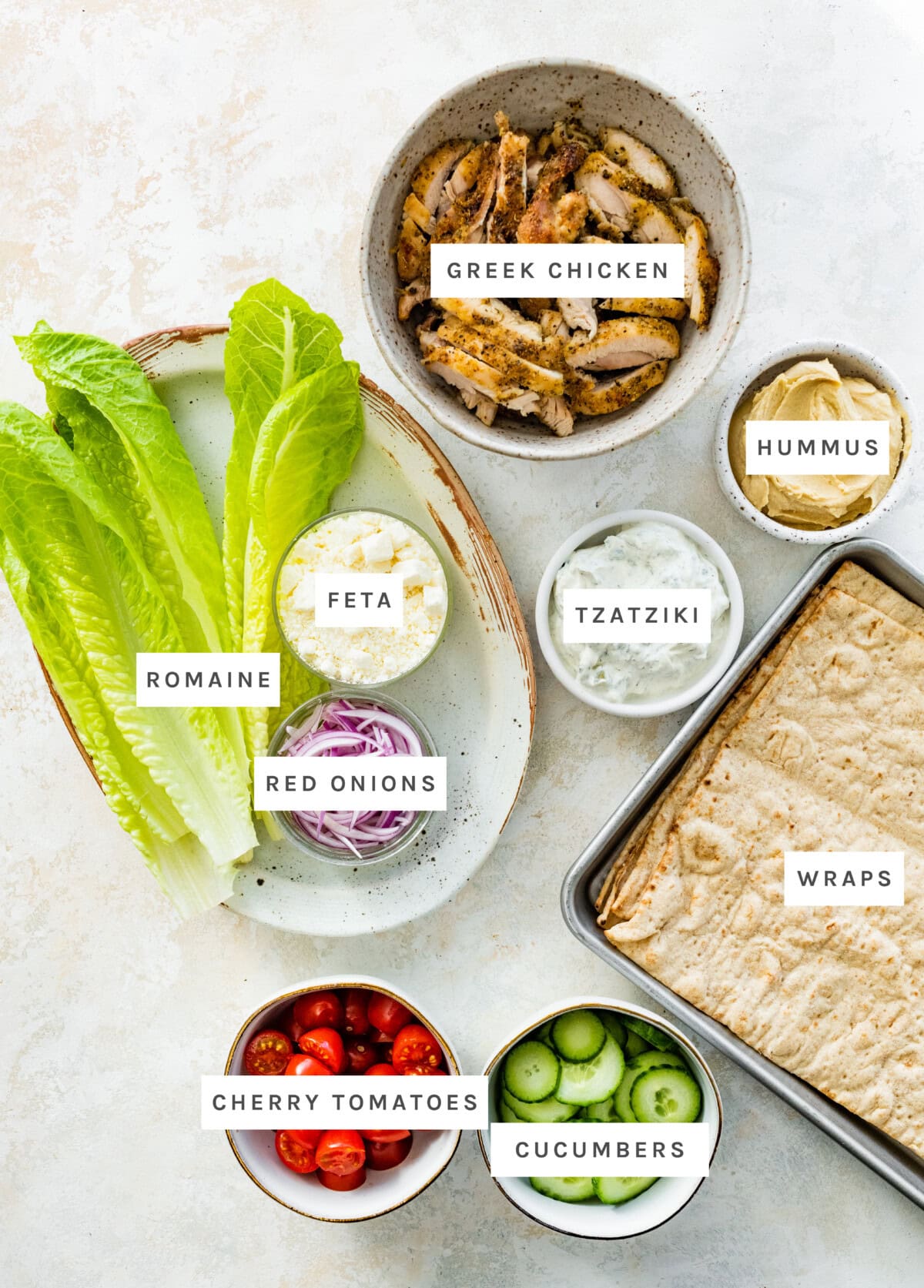 Ingredients measured out to make Greek Chicken Wraps: Greek chicken, hummus, tzatziki, wraps, romaine lettuce, feta cheese, red onions, cherry tomatoes, cucumbers.
