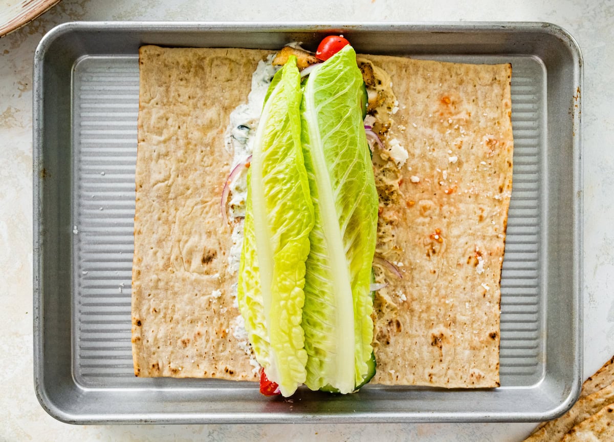A piece of lavash bread filled with hummus, tzatziki, red onion, tomato, Greek chicken, cucumber, feta and romaine lettuce.