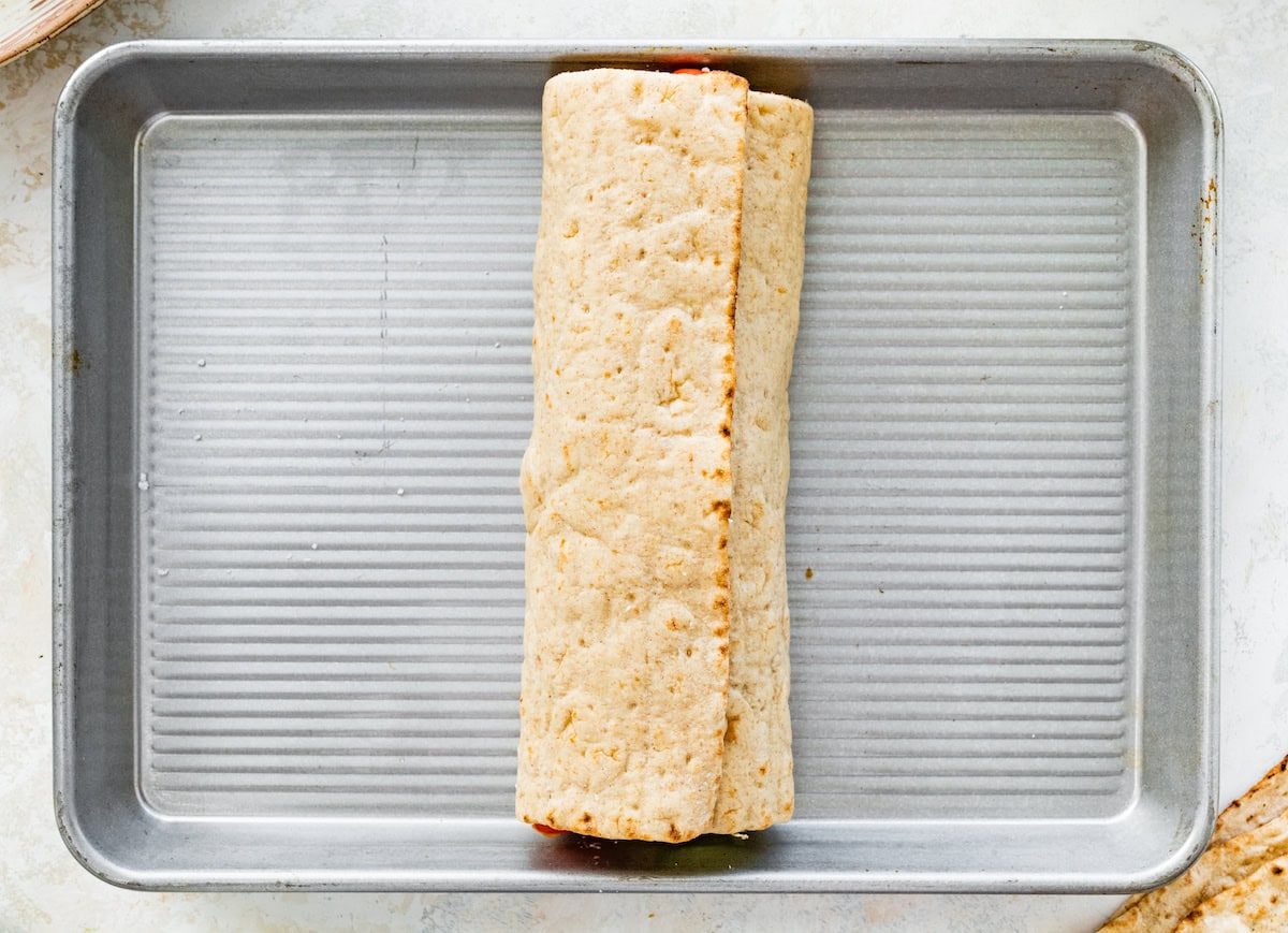 Lavash bread wrapped up to make a Greek chicken wrap. The wrap is on a silver baking sheet.