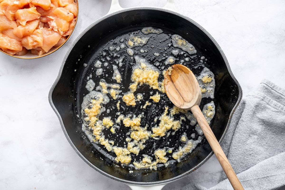 A cast iron skillet with melted butter and minced garlic sizzling, with a wooden spoon stirring the mixture. A bowl of raw chicken sits beside the skillet.