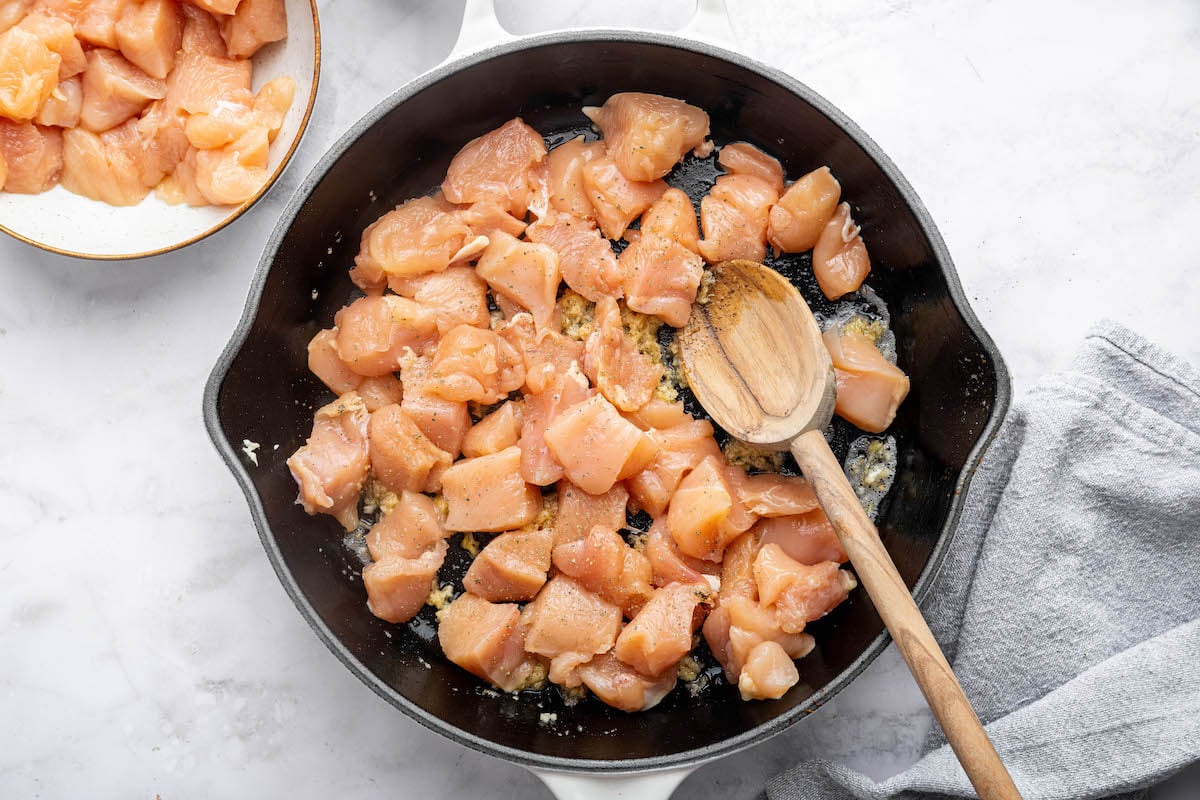 A cast iron skillet with diced raw chicken cooking over minced garlic and butter, being stirred with a wooden spoon.