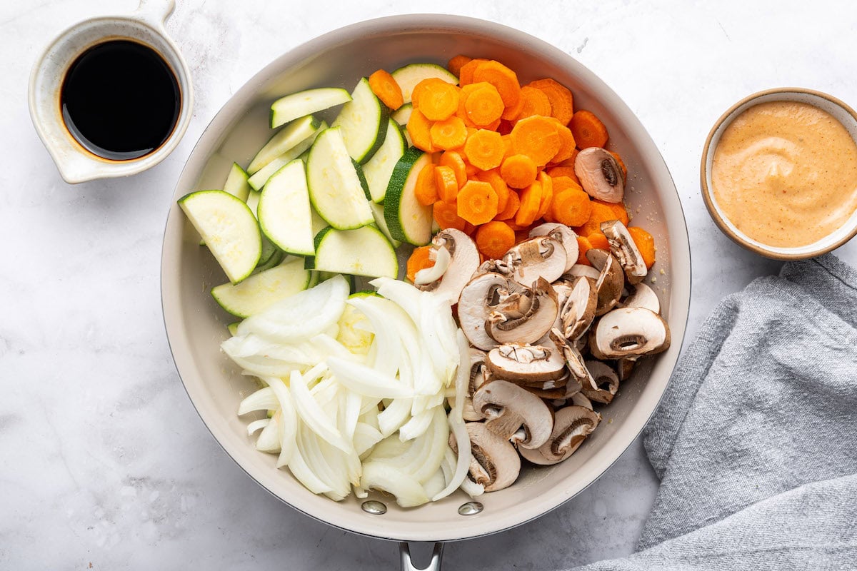A white skillet filled with fresh, uncooked vegetables—sliced zucchini, carrots, mushrooms, and onions—arranged neatly, with a bowl of sauce nearby.
