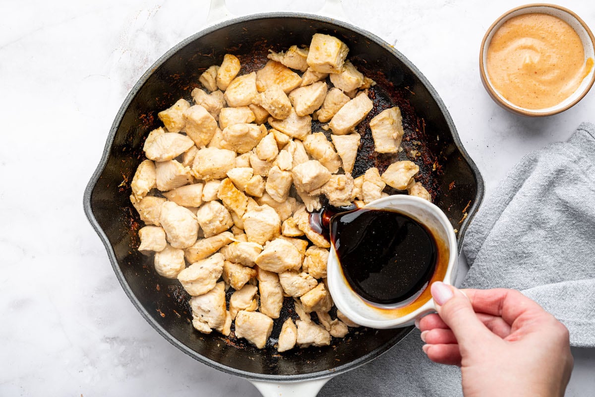 A cast iron skillet with golden-brown cooked chicken, with a hand pouring a dark soy sauce-based marinade over the chicken.