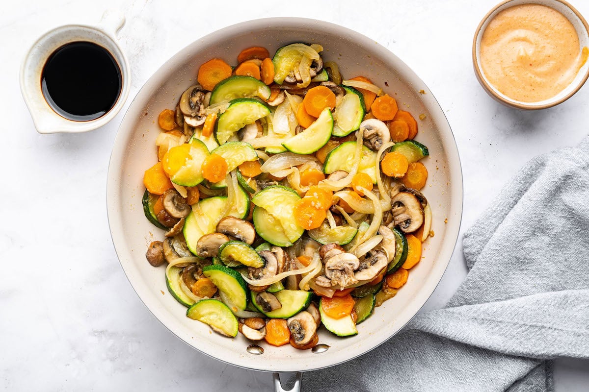 A white skillet with stir-fried vegetables, including zucchini, carrots, mushrooms, and onions, lightly coated in sauce.