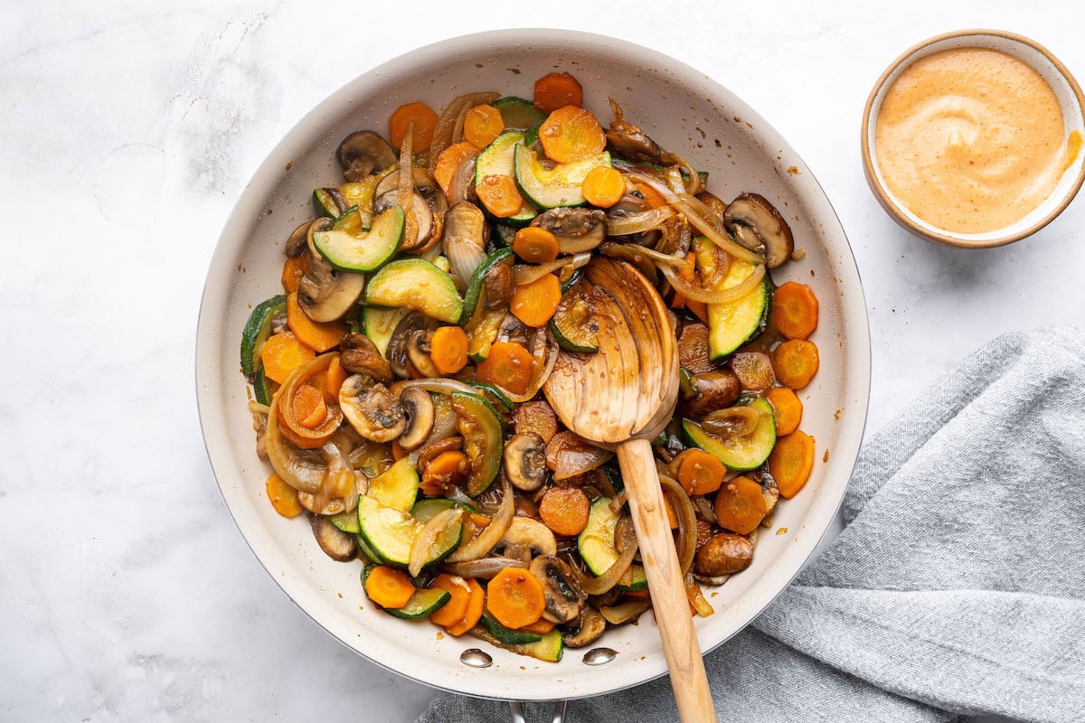 A white skillet with stir-fried vegetables, showing a wooden spoon stirring the cooked zucchini, carrots, mushrooms, and onions.