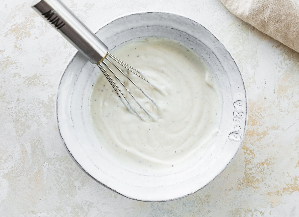 A white ceramic bowl filled with a creamy Greek yogurt sauce being whisked with a metal whisk.