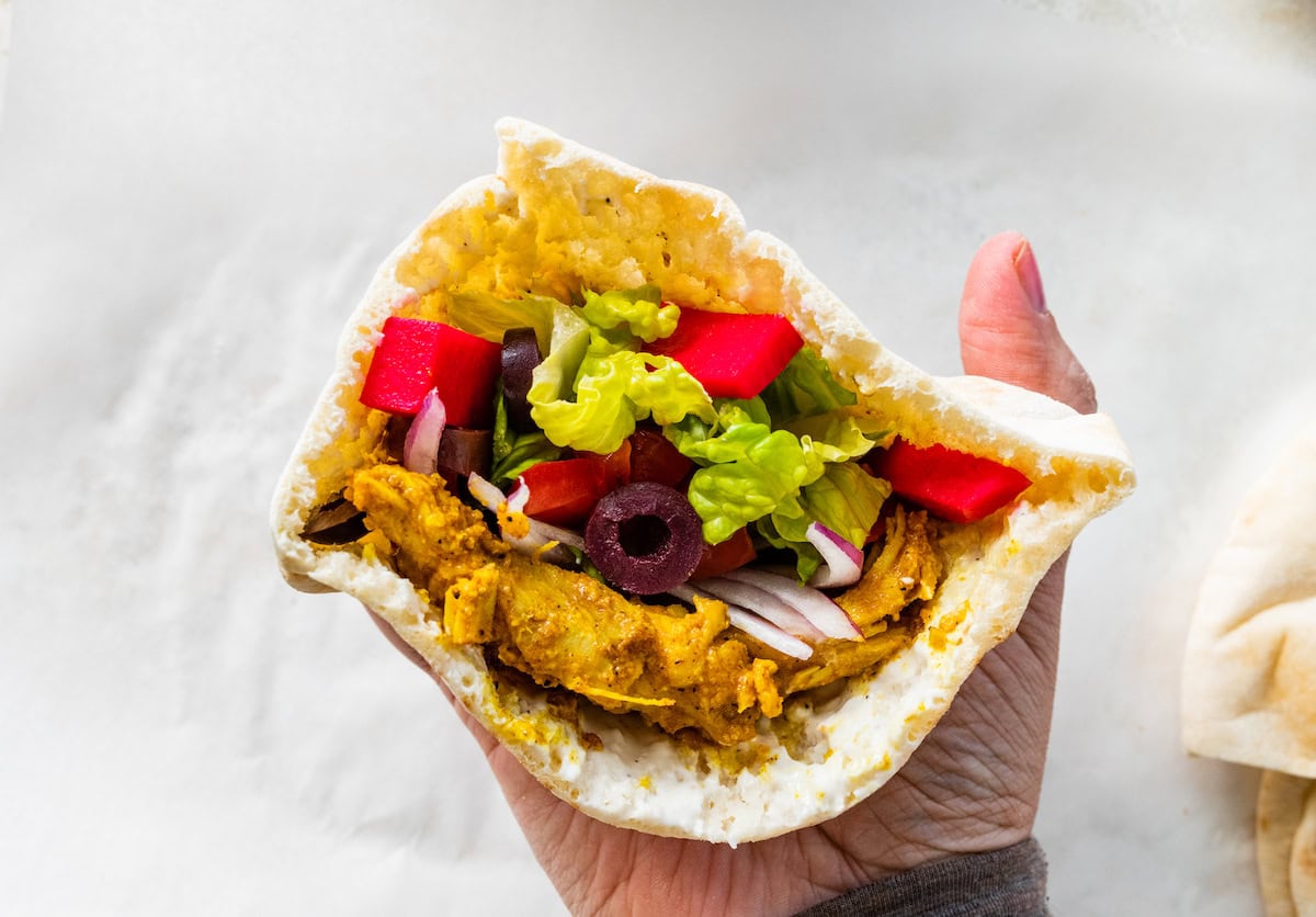 A hand holding a chicken shawarma pita pocket filled with lettuce, tomatoes, pickled turnips, red onion, and Kalamata olives without sauce.