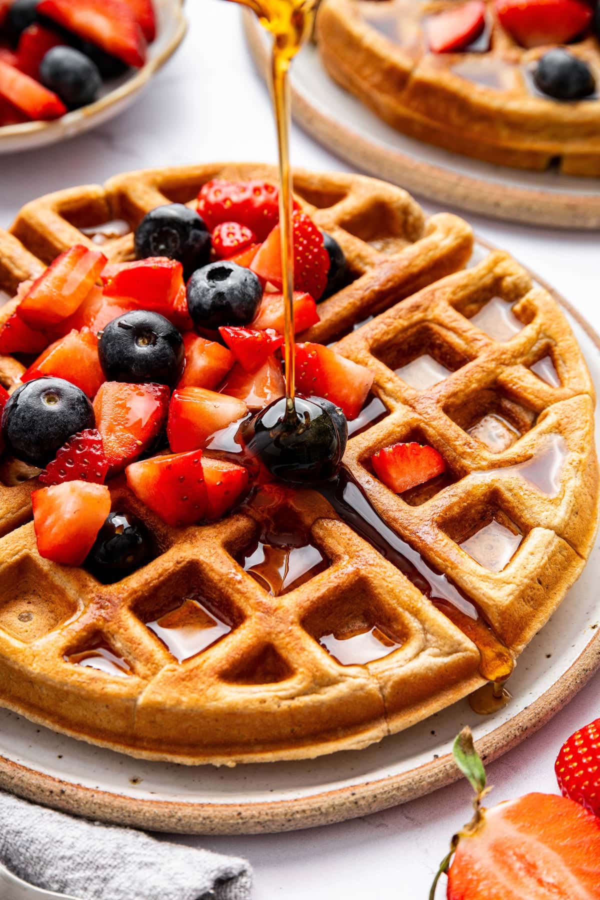 Golden brown protein waffles topped with fresh strawberries and blueberries, as a stream of maple syrup drizzles over the top.