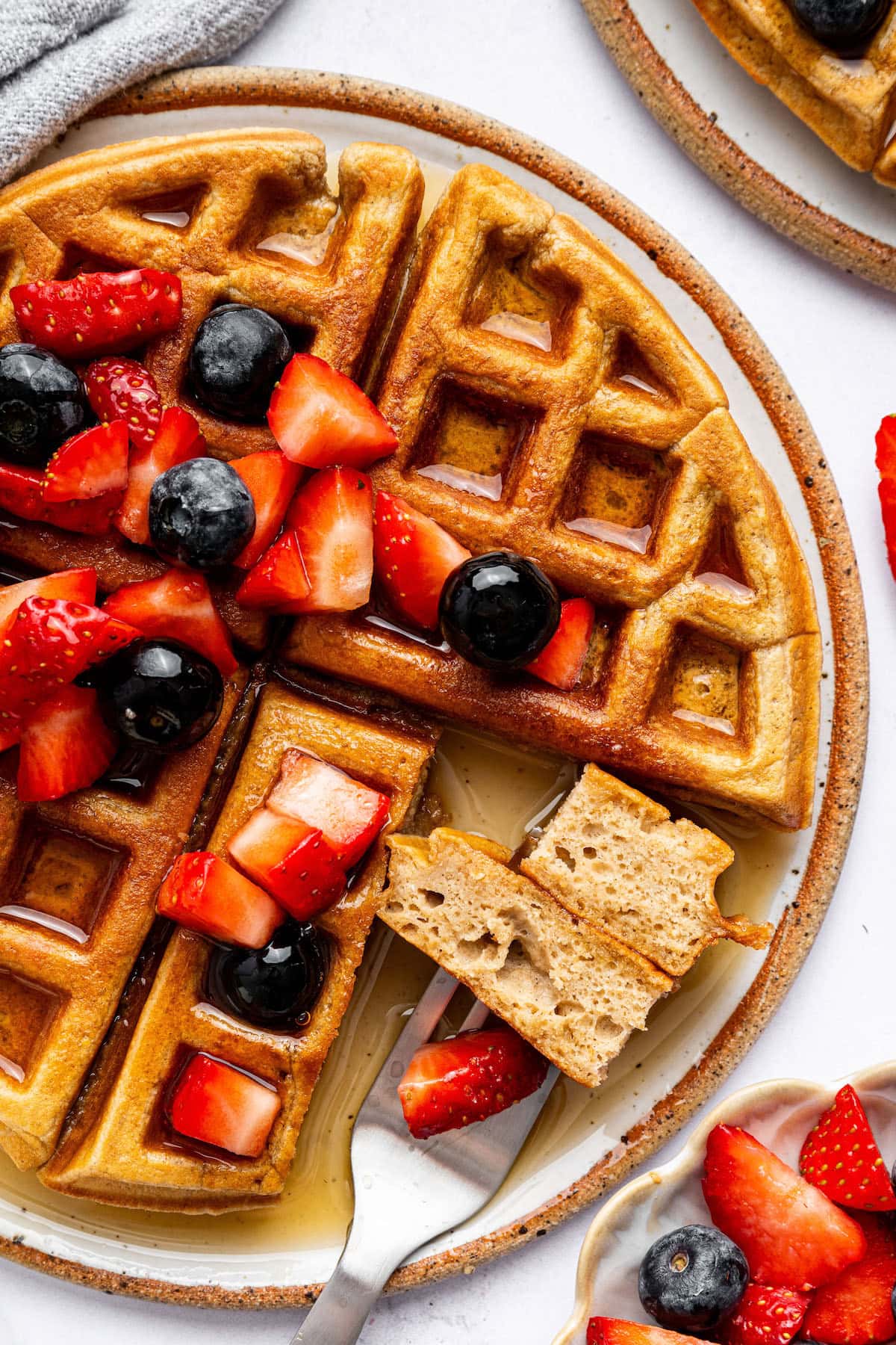 Golden brown protein waffles on a plate, topped with fresh strawberries and blueberries, drizzled with syrup, and served with a side of fruit.