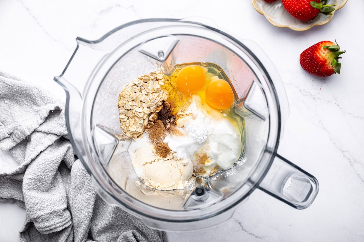 A blender filled with ingredients for protein waffles, including rolled oats, eggs, yogurt, baking powder, and spices.