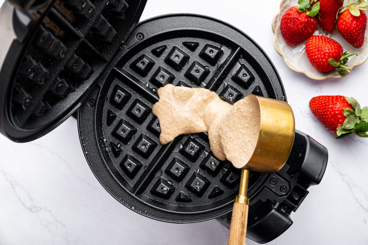 A scoop of thick protein waffle batter being poured into a preheated waffle iron, with fresh strawberries in the background.