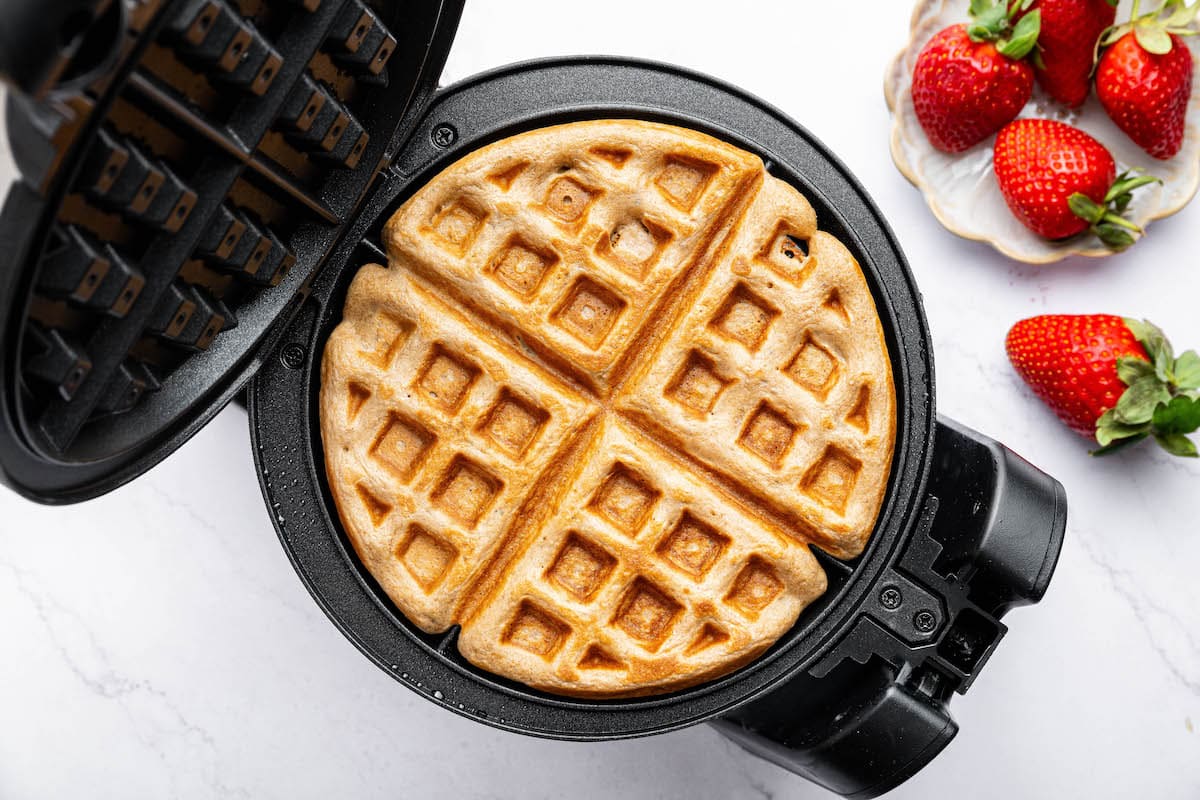 A freshly cooked protein waffle inside a waffle maker, golden brown and crispy, with a plate of strawberries nearby.