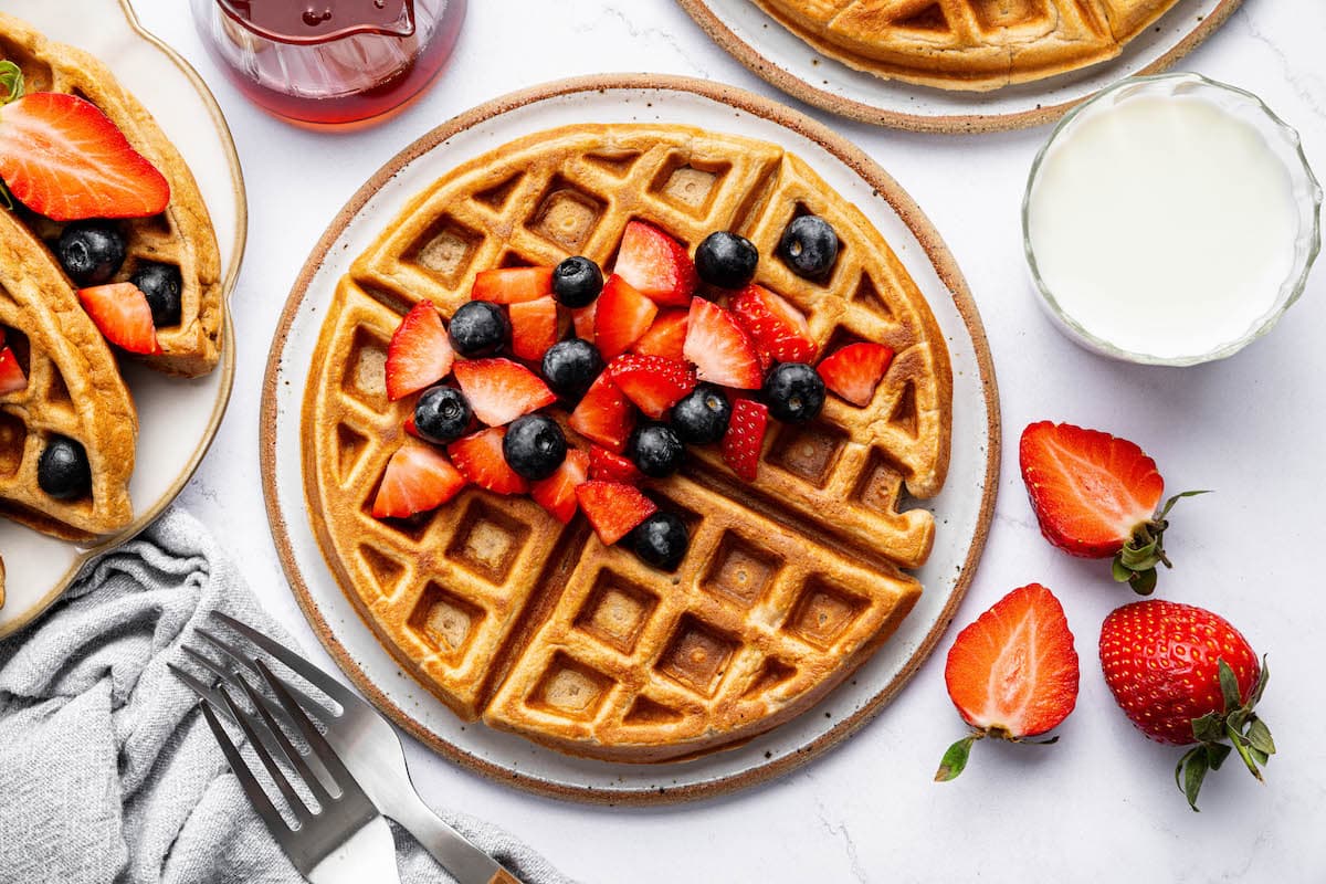 Plated protein waffles topped with sliced strawberries and blueberries, served with a glass of milk and a small jar of maple syrup.