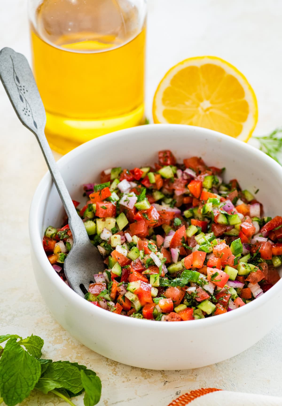 Shirazi Salad in a small white bowl served with a silver fork. There is a lemon half and a bottle of olive olive in the background.