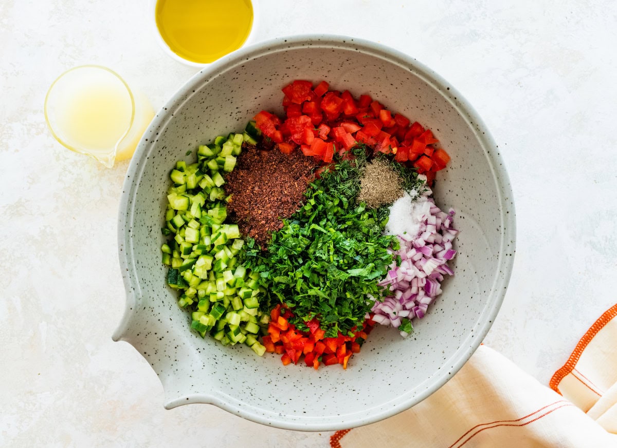 The ingredients for the Shirazi Salad in a mixing bowl: cucumbers, tomatoes, red onion, bell pepper, sumac, salt, pepper and fresh parsley, mint and dill.