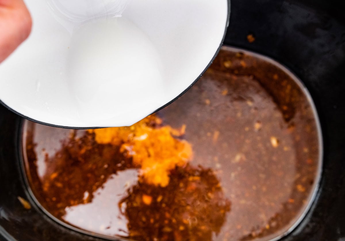 A woman's hand pouring the arrowroot powder mixture into the teriyaki sauce in the slow cooker.