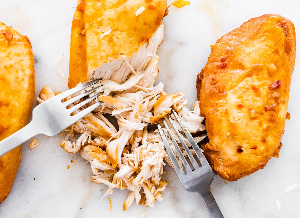 Three teriyaki chicken breasts being shredded with two forks.