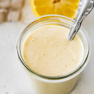 A creamy tahini dressing in a glass jar with a metal spoon and a halved lemon in the background.