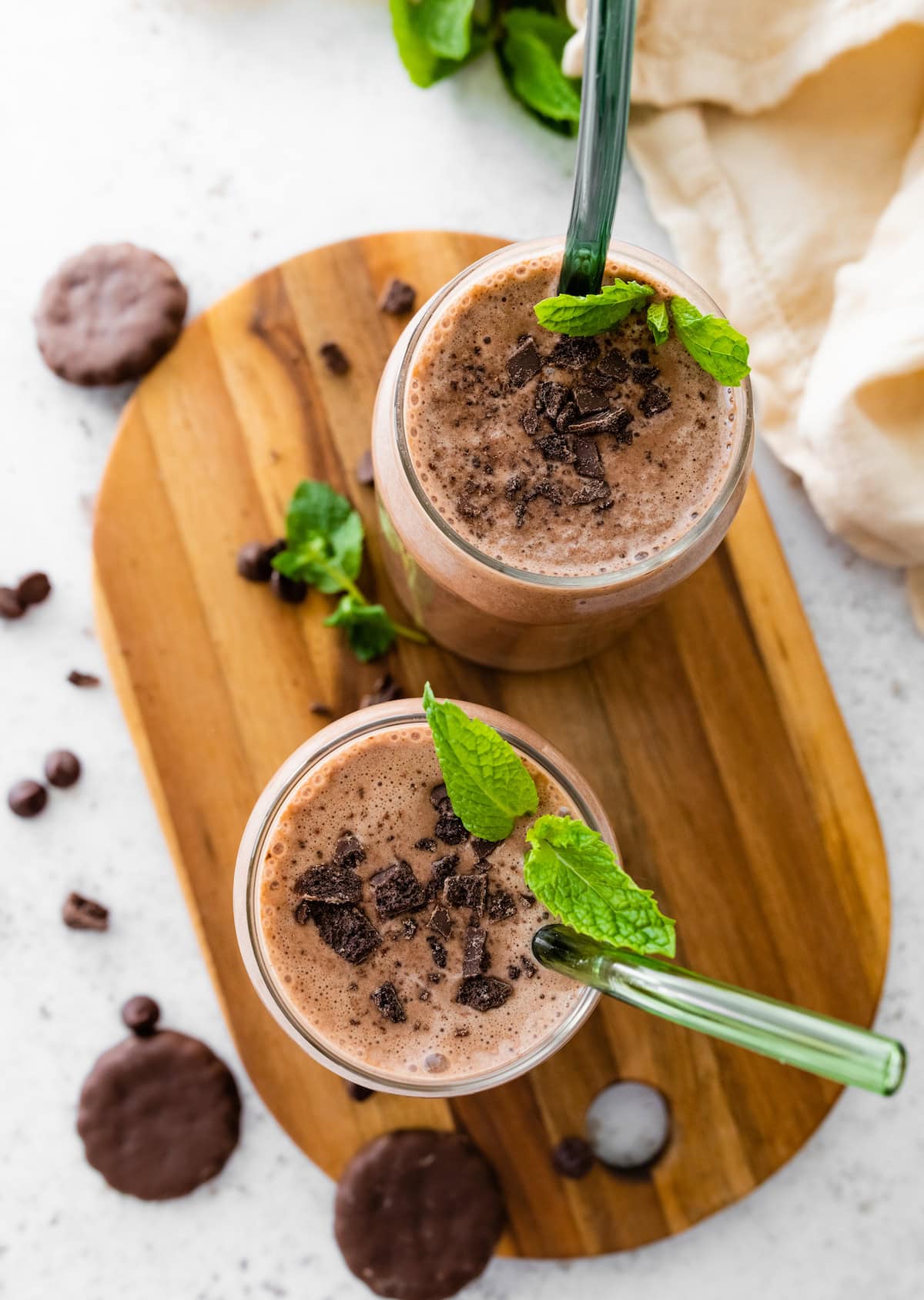 Two Thin Mint protein shakes in glasses topped with crushed Thin Mint cookies and fresh mint. There are green straws in both glasses.