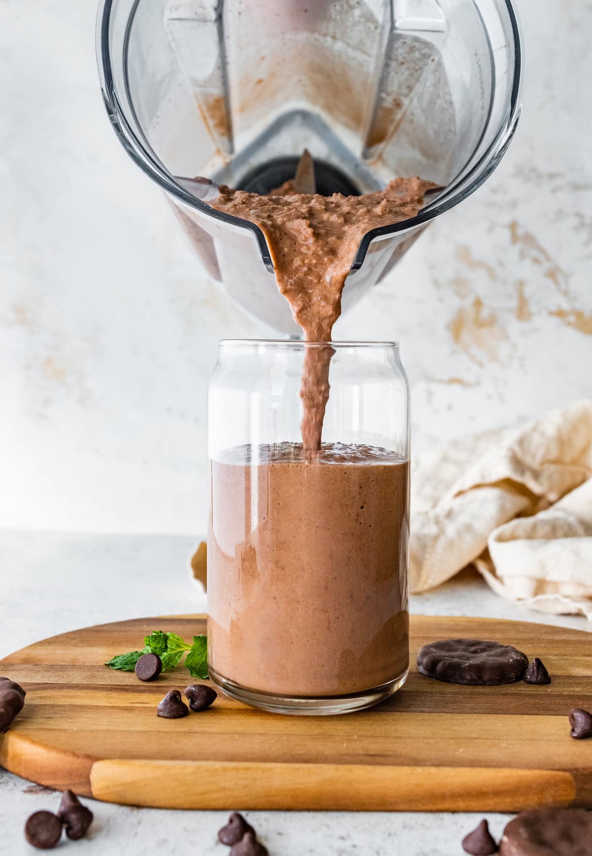 A mint chocolate protein shake being poured into a glass from a Vitamix blender. The glass is sitting on a wooden cutting board.