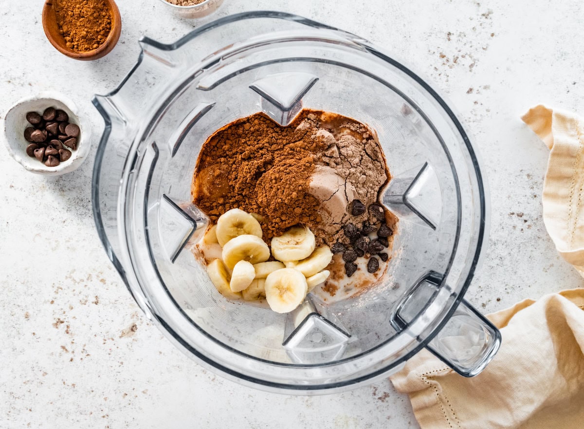 The ingredients for the Thin Mint protein shake in the base of a Vitamix blender: frozen banana, cocoa powder, chocolate protein powder, peppermint extract, almond milk and chocolate chips.