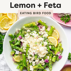 This image showcases a fresh asparagus salad with lemon and feta cheese. The dish includes vibrant green asparagus, crumbled feta cheese, red onions, and fresh parsley, garnished with lemon slices. A small bowl of dressing is placed nearby, indicating it is likely drizzled over the salad for added flavor. The scene is complemented by a pink glass of water, a small dish of extra feta, and a plate of lemon slices, all presented on a light, bright background with a soft, airy aesthetic.