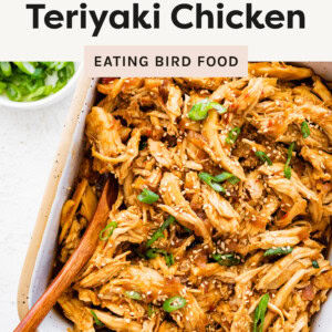 Overhead shot of slow cooker teriyaki chicken, shredded and coated in a glossy, savory-sweet sauce. The dish is garnished with sesame seeds and sliced green onions, served in a square ceramic dish with a wooden spoon. A small bowl of extra green onions is placed beside it.