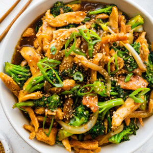 Chicken and broccoli stir fry in a white bowl, garnished with sesame seeds and green onions, with wooden chopsticks beside it.