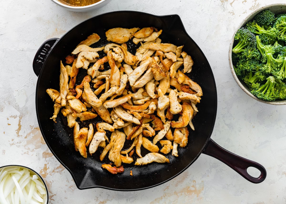 Cooked chicken in a cast iron skillet next to a bowl of raw broccoli florets and sliced onions.