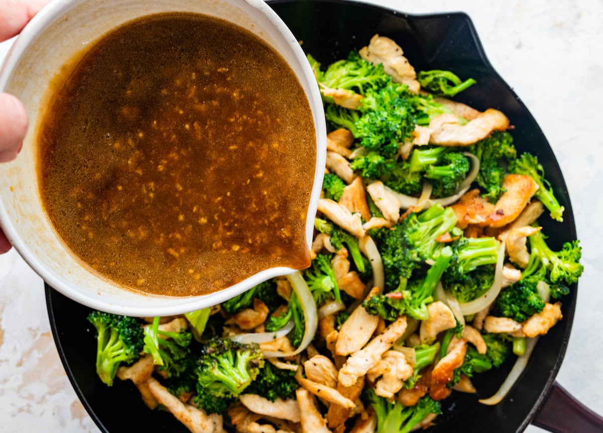 A woman's hand pouring the stir fry sauce over the chicken and broccoli stir fry in a cast iron skillet.