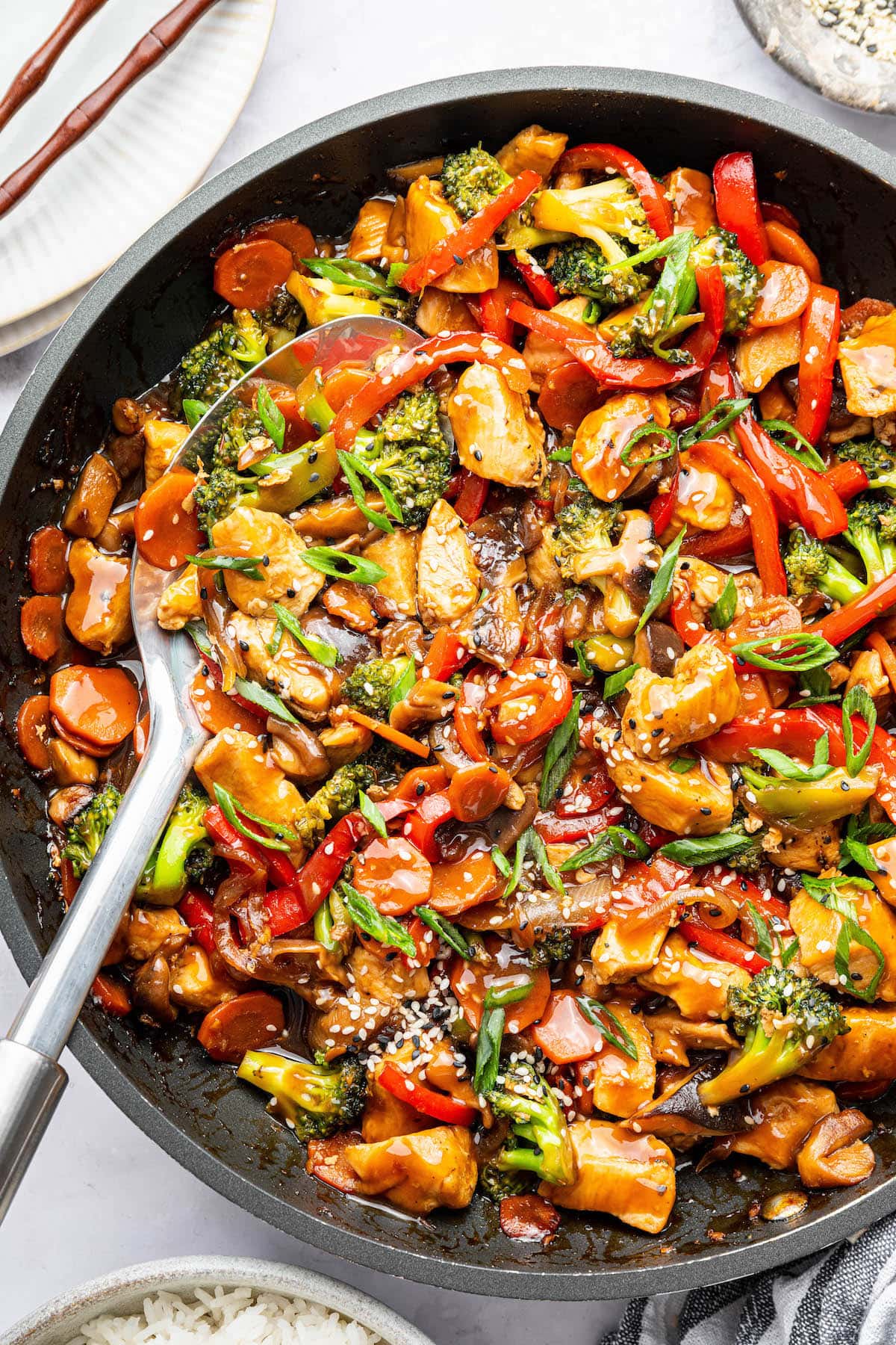 Chicken stir fry with colorful vegetables and tender chicken in a skillet, topped with sesame seeds and green onions. There is a silver serving spoon in the skillet.
