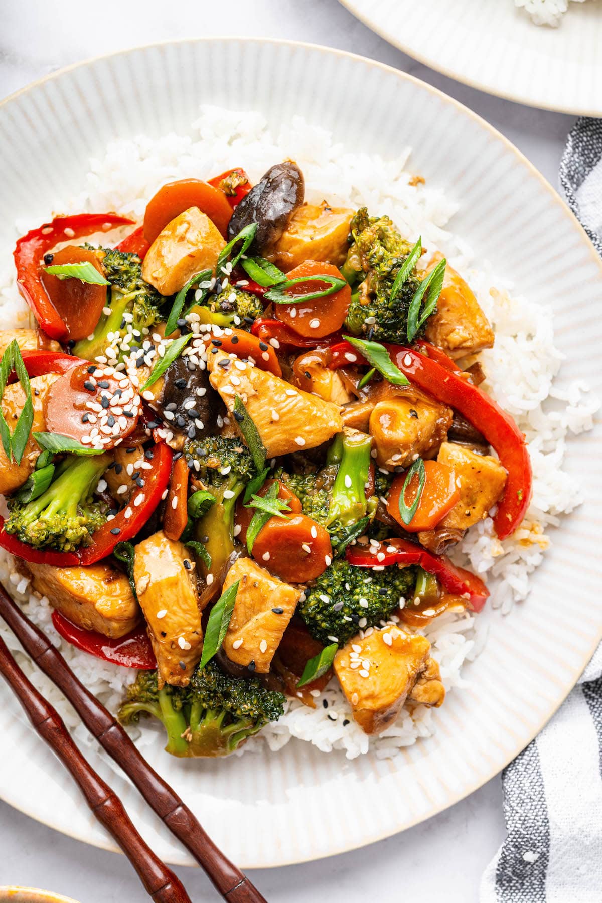 Chicken stir fry with colorful vegetables and tender chicken served on a white plate with white rice, topped with sesame seeds and green onions.