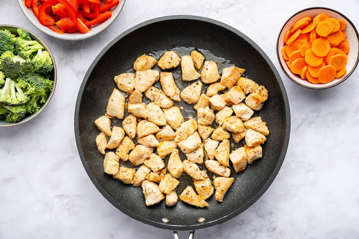 Cooked chicken chunks in a skillet. There are three bowls next to the skillet filled with broccoli florets, chopped carrots and red bell pepper.