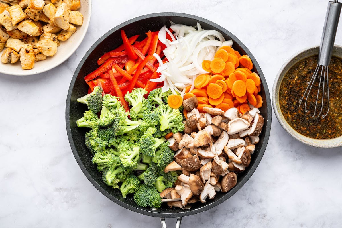 Fresh veggies in a skillet: red bell pepper, yellow onion, carrots, broccoli, and mushrooms, with cooked chicken and stir fry sauce beside the skillet.