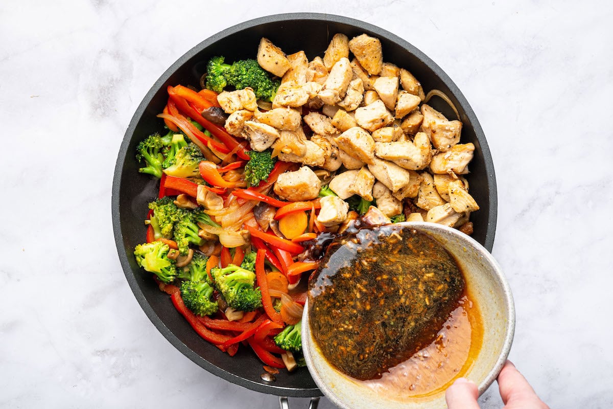 A woman's hand pouring the stir fry sauce over the cooked chicken and cooked vegetables in a skillet.