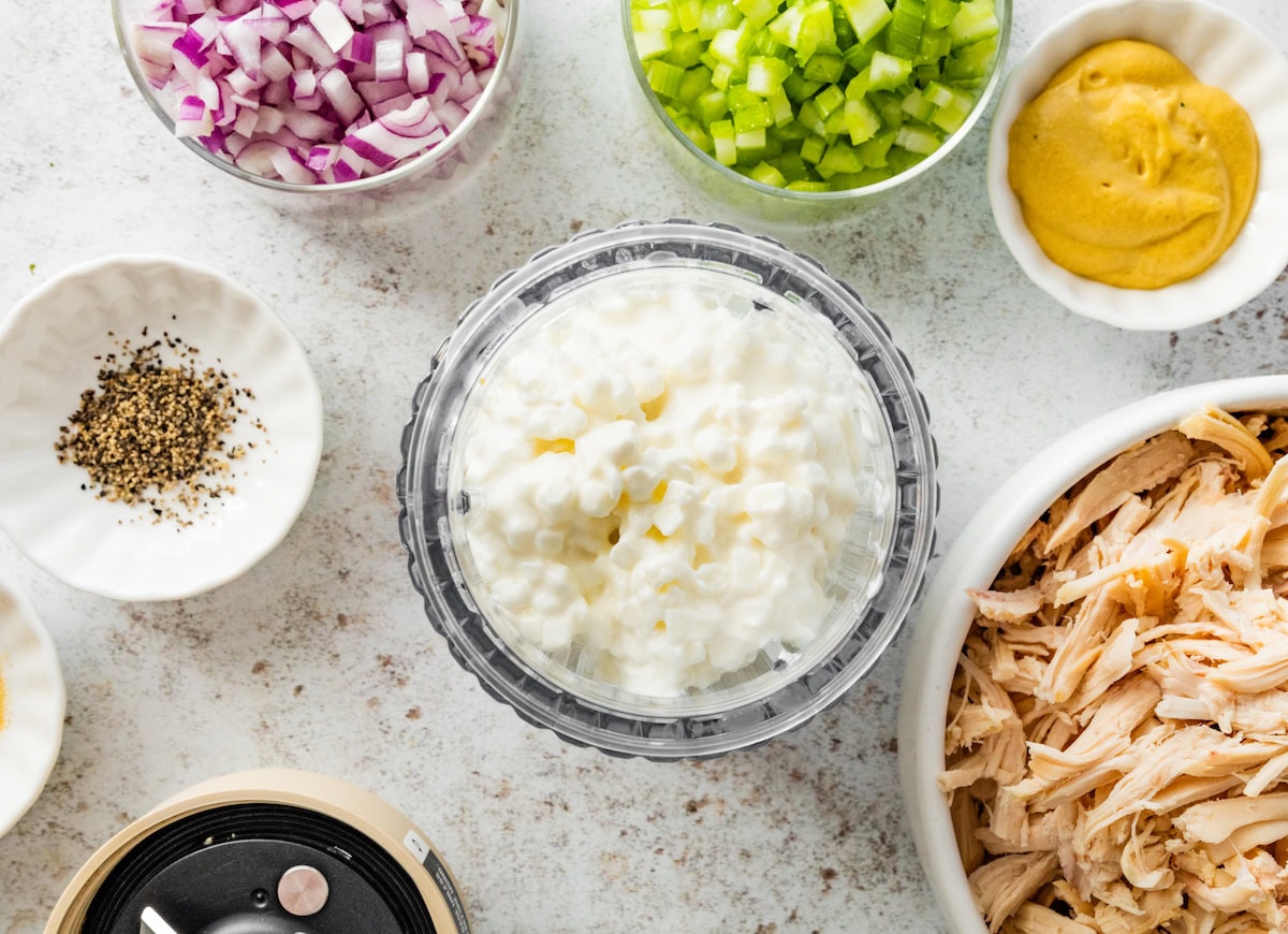 Cottage cheese in a small blender. There are multiple bowls next to it with shredded chicken, celery, red onion, pepper and dijon mustard.