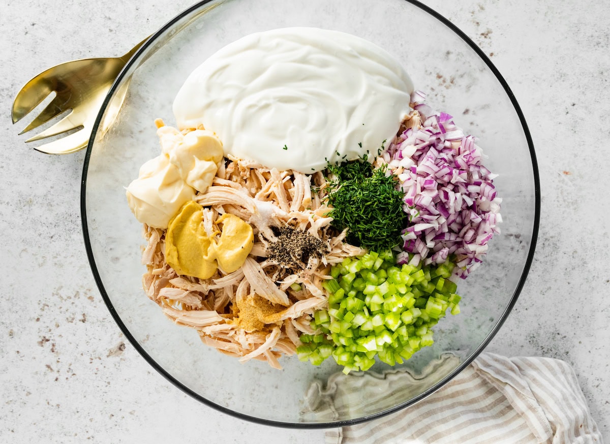 The ingredients for the cottage cheese chicken salad in a glass mixing bowl: blended cottage cheese, shredded chicken, dijon mustard, red onion, celery, fresh dill, mayo, salt and pepper.