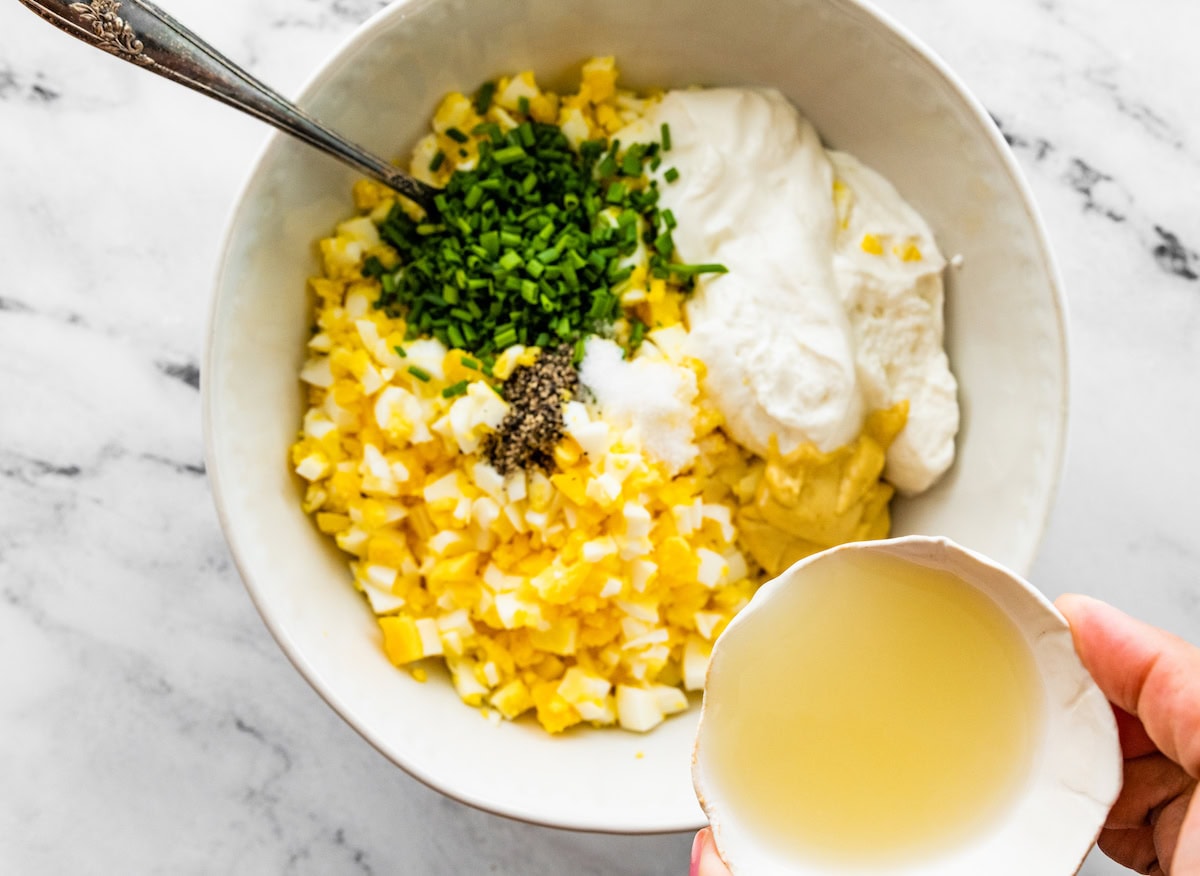 Chopped hard boiled eggs in a white ceramic bowl with blended cottage cheese, dijon mustard, chives, salt and pepper. A woman's hand is getting ready to pour lemon juice into the bowl.