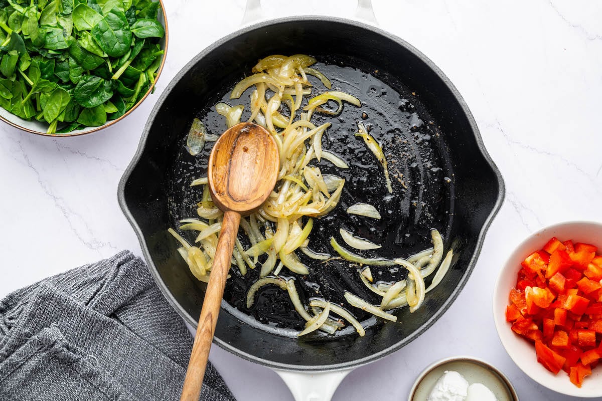 Caramelized onions cooking in a white cast iron skillet with a wooden spoon.