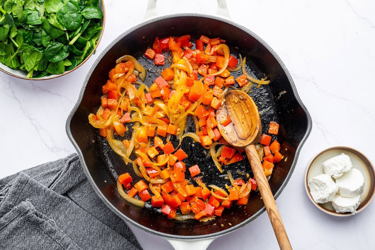 Red bell peppers being sautéed in a white cast iron skillet with a wooden spoon.