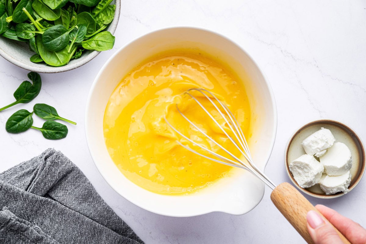 A woman's hand whisking the egg mixture for the frittata recipe in a white mixing bowl.