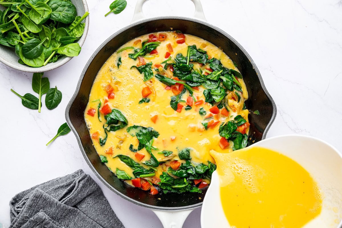 The egg mixture being poured over the cooked veggies in a cast iron skillet for the frittata recipe.